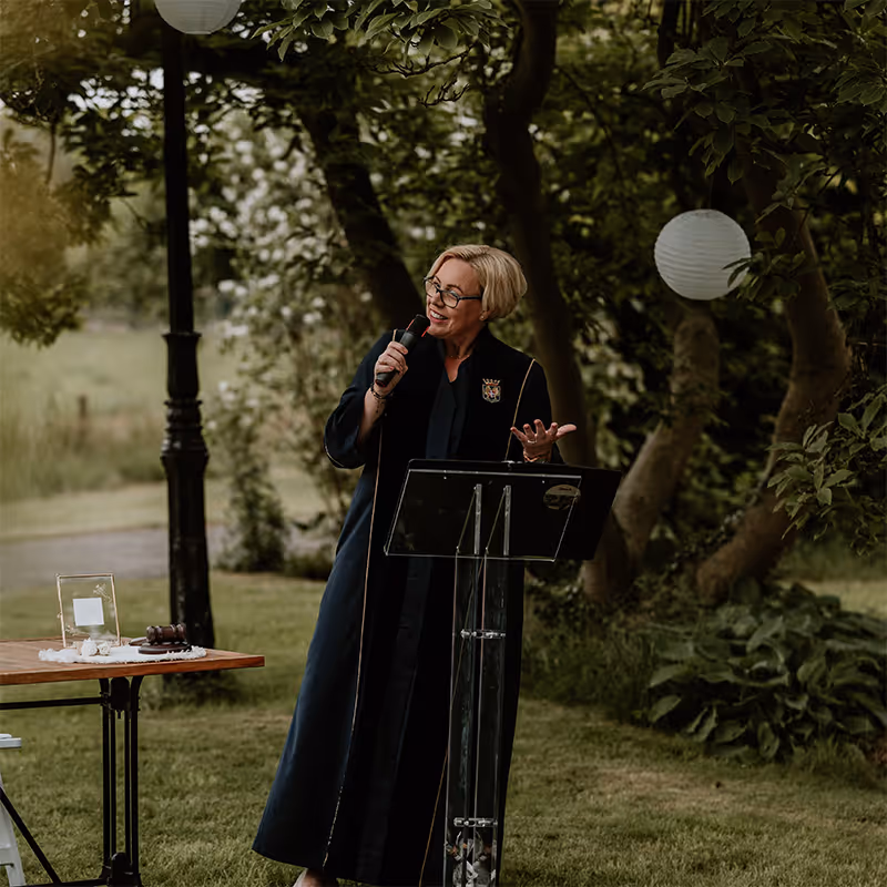 Trouwambtenaar karin aan het spreken op een ceremonie in een mooie tuin in groene toga