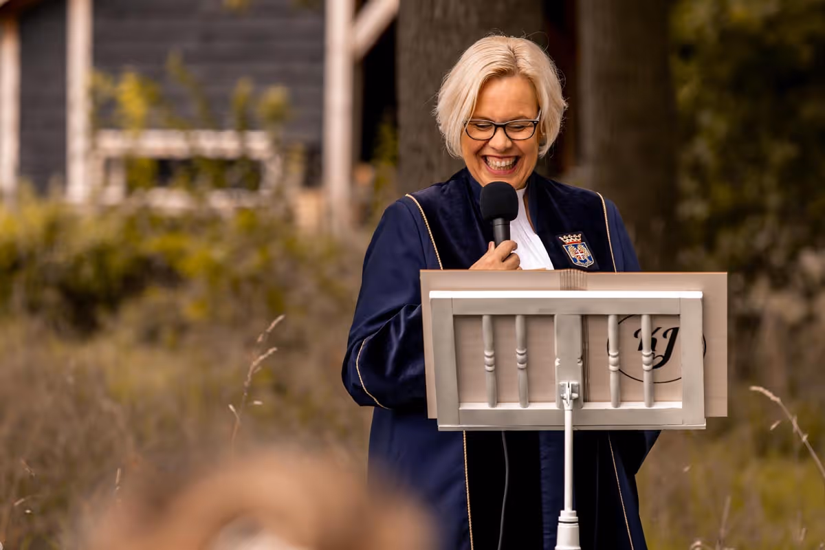 close up foto van trouwambtenaar karin acahter katheder met microfoon in de hand, ze heeft een blauwe toga aan en lacht voluit