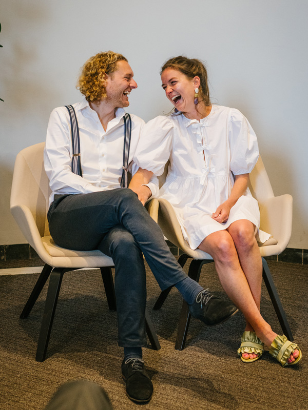 Bruidspaar zit hand in hand naast elkaar bij de ceremonie  van een huwelijk in Twente en lachen uitbundig naar elkaar