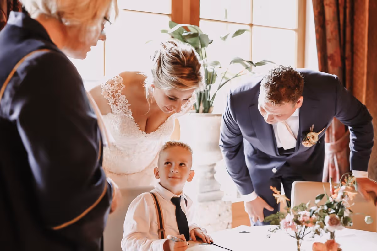jongente tekent kinderakte, trouwsmbtenaar karin staat er naast en het jongente kijkt omhoog naar Karin, het bruispaar staat gebogen achter het jongentje en kijkt mee naar de akte