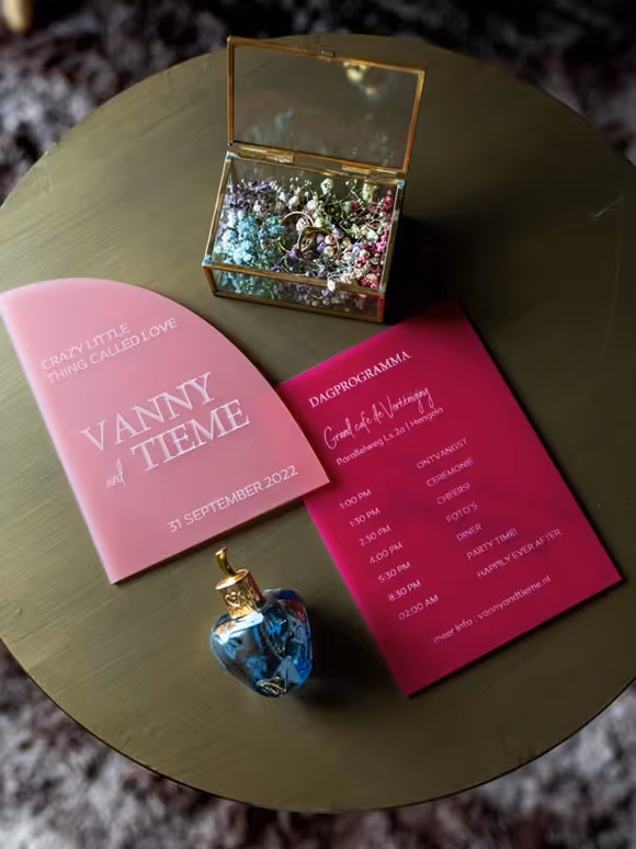 flatlay van roze acryl uitnodigingen en ringen in gouden glazen ringdoosje met gekleurd gipskruid op een gouden tafeltje